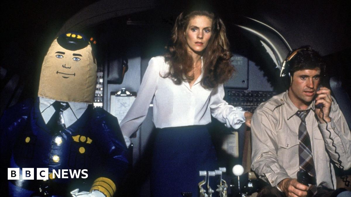 A colour mid-shot, a publicity still from the film Airplane!. It shows a jet airliner cockpit set, with seated on the left of frame the inflatable autopilot character ‘Otto’; standing in the centre a woman in her mid-20s in the white blouse and dark blue skirt uniform of an airline cabin crew member; and on the right of frame, sitting, a man in his early thirties with headphones on, in light grey shirt and greyscale tartan tie, operating the controls and holding a radio set microphone.