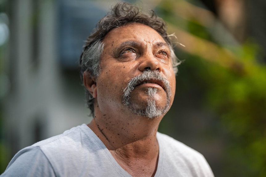 Tanned man, brown hair, gray sideburns and moustache goatee, white t-shirt looking up to the sky, greenery blurred background