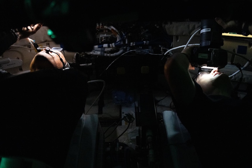 two astronauts sit in darkness pearing up into camera viewfinders, their faces iluminated 