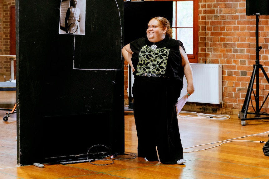 Megan Wilding, an Indigenous woman in her mid-30s, smiles brightly in a brick-walled rehearsal space, one hand on her hip.