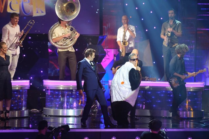 A TV still of Mikey Robins, 40s, in a penguin-style costume and sunglasses, with Paul McDermott, a live band playing behind.