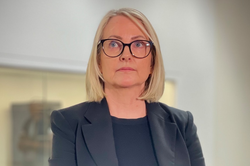 A woman with blonde hair and black glasses standing in front of microphones