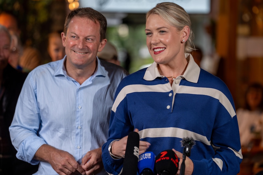 Josh Teague is smiling next to Liberal leader Ashton Hurn as she stands behind a group of media microphones