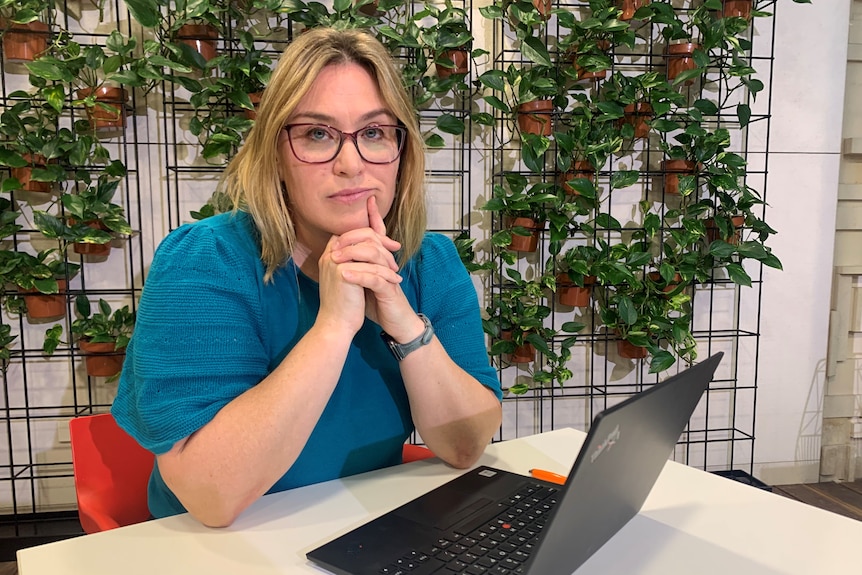 A woman in a blue shirt and glasses sits at a desk with a laptop.