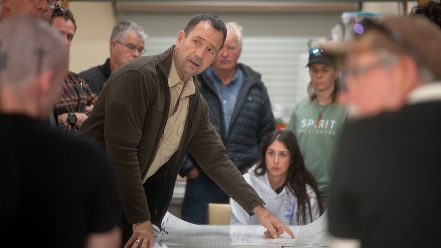A man in a brown shirt and sweater leans over a laminated map, pointing. A crowd of people stand around him
