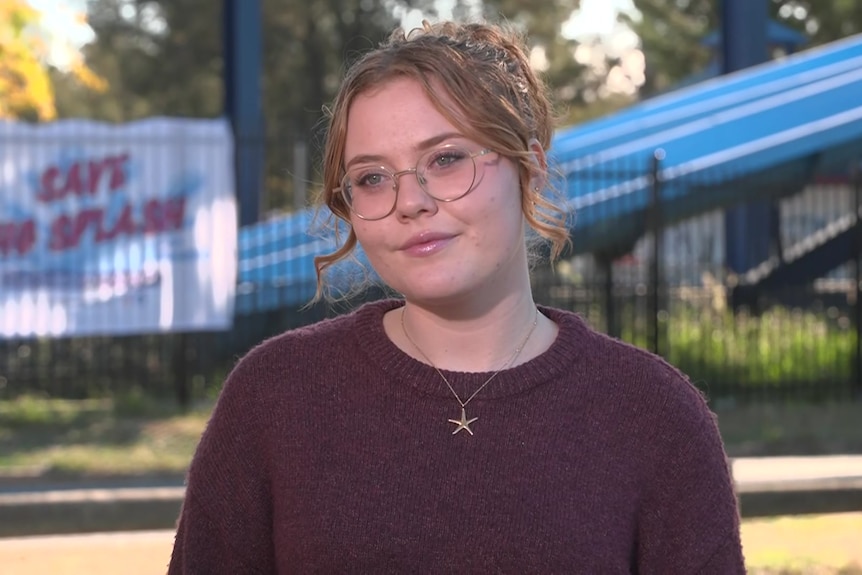 A young woman with long light brown hair stands in front of a derelict water park.