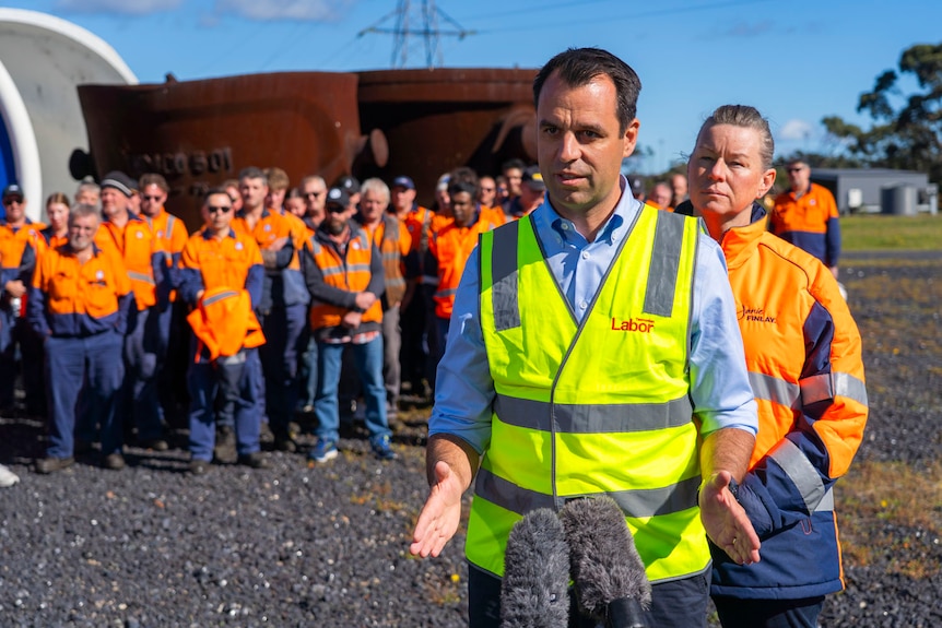 man in fluro yellow vest stands in front of workers in orange