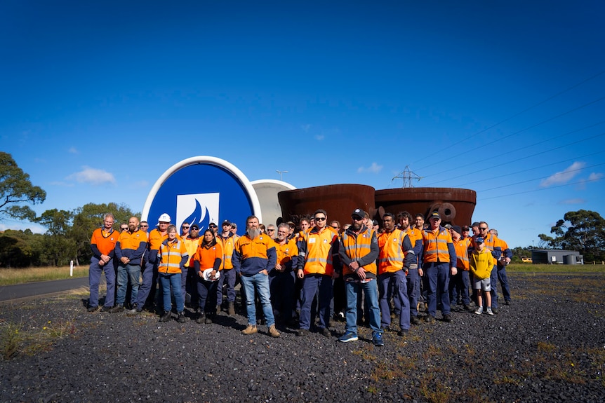 men and women in orange high vis outside a blue logo and a rusted sculpture
