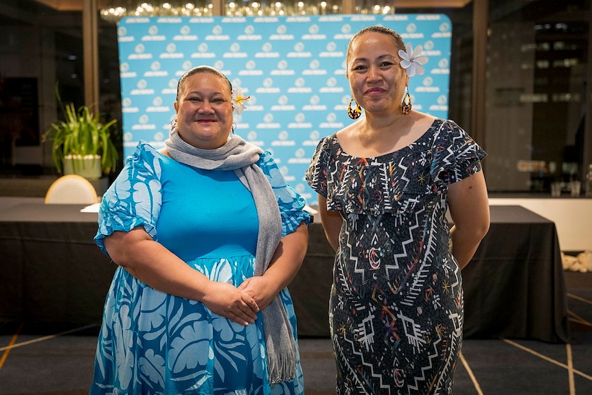 Two women, each with a flower behind their left ear, smile for the camera.