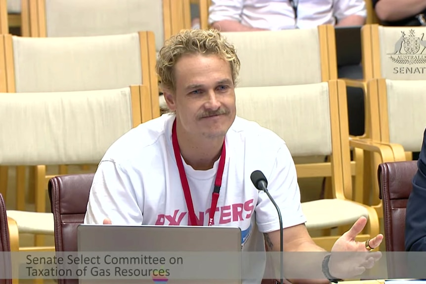 A man in a white tshirt with blonde hair sits in front of a microphone