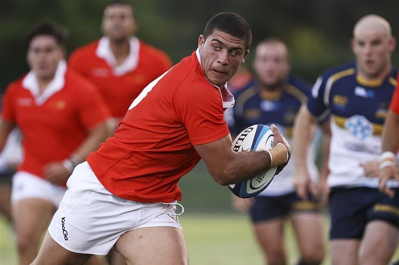 A man in a red jersey holds a rugby ball as he runs.