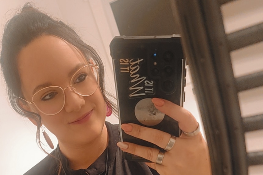 A dark-haired woman in glasses smiles as she takes a selfie in a bathroom.