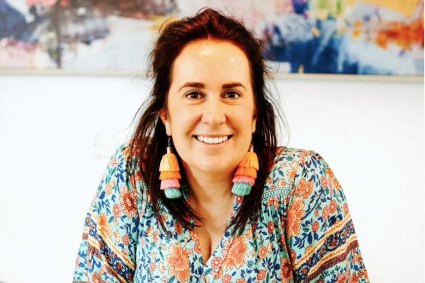 A woman sits a table with colourful earrings and shirt, with painting in the background.