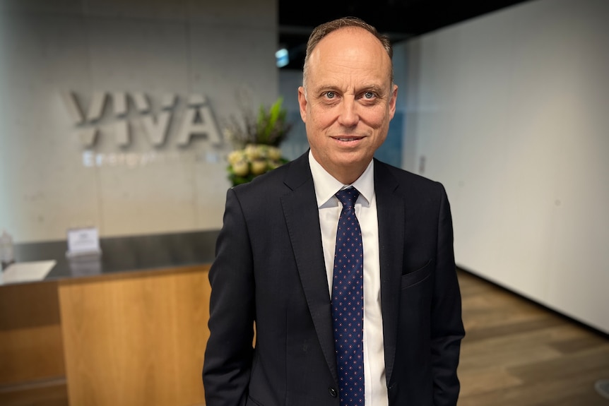 Scott is slightly smiling, wears a black suit and blue tie with pink polka dots, is balding, Caucasian, Viva on wall behind.