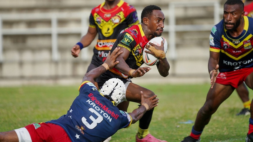 A PNG man holding a white rugby league ball is running through a missed tackle by a man in white head gear.