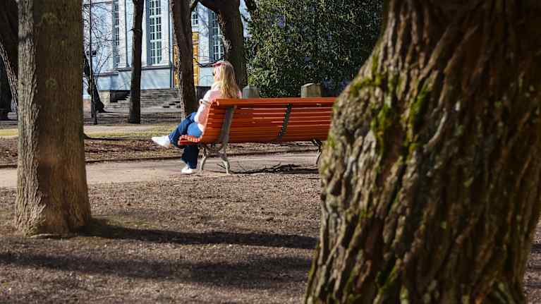 Woman wearing jeans and a pink jacket sitting on a park bench, surrounded by trees and an old house can be seen in the background.