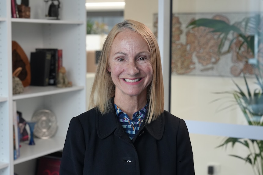 A smiling woman with straight, blonde hair,  standing in front of a painting.