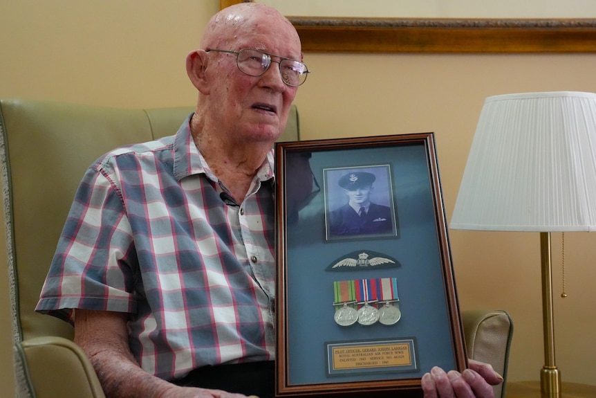Elderly white man in a plaid button-up shirt, holding a wooden photo frame, with image at the top, and service medals below.
