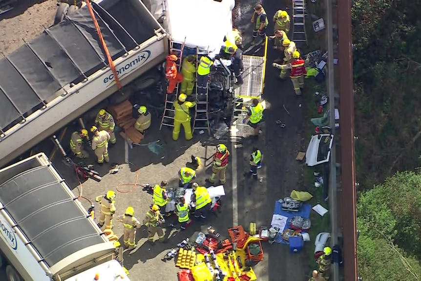 Dozens of emergency workers, includings ome on ladders and scaffolding crowd around the crashed cabin of a truck on a sunny day.
