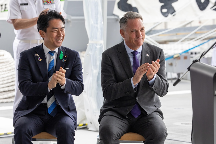 Two men sit side by side, smiling and clapping, on the deck of a frigate