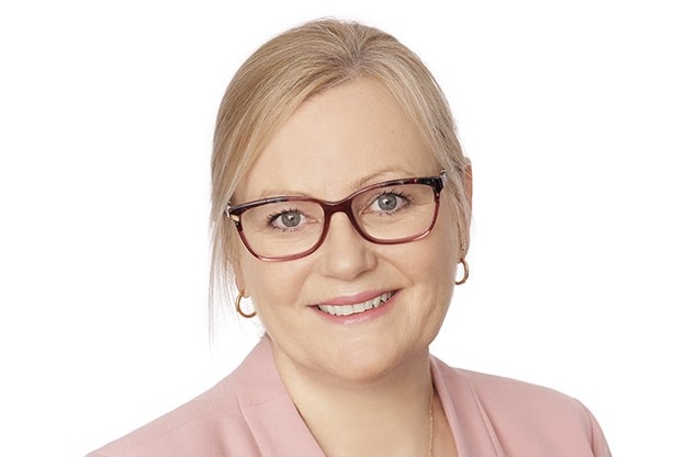A blonde middle-aged woman with glasses, small hoop earrings and hair tied back looks at the camera smiling