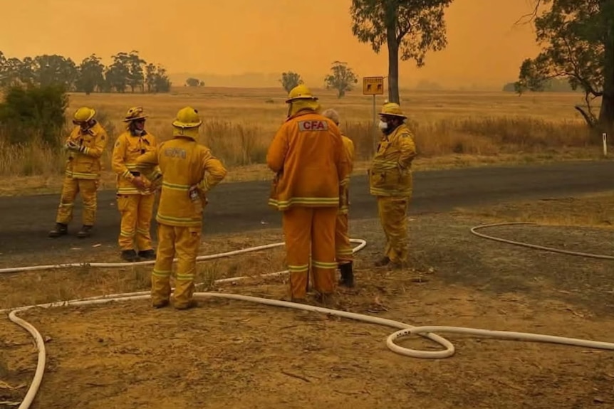 A small group of firefighters gathered in a bush setting. The sky is yellow with smoke haze. 
