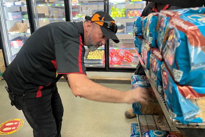 A man inside a small grocery store, he is inspecting stock lined up on a shelf.