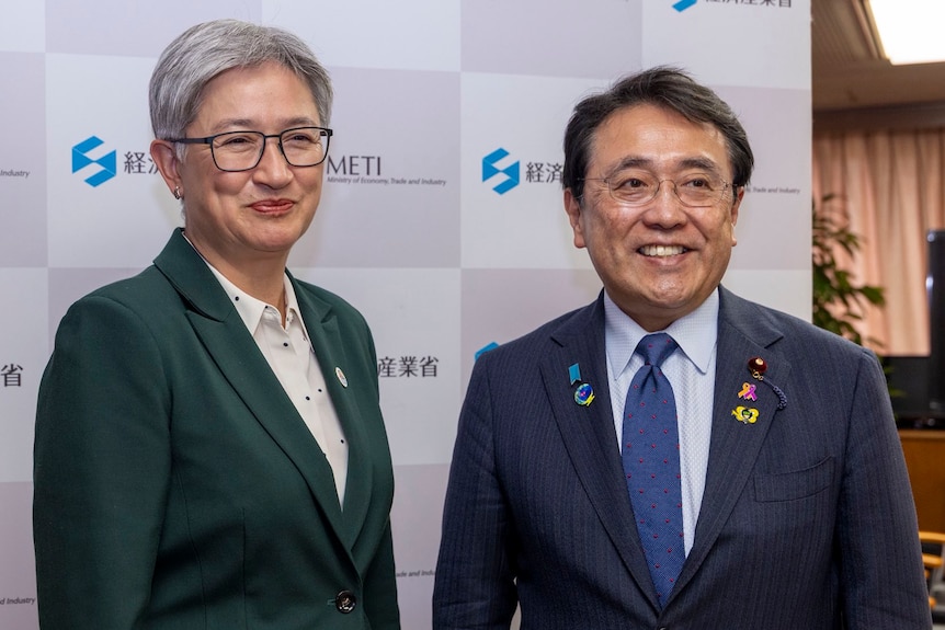 A woman in a green blazer stands smiling next to a man in blue blazer and tie