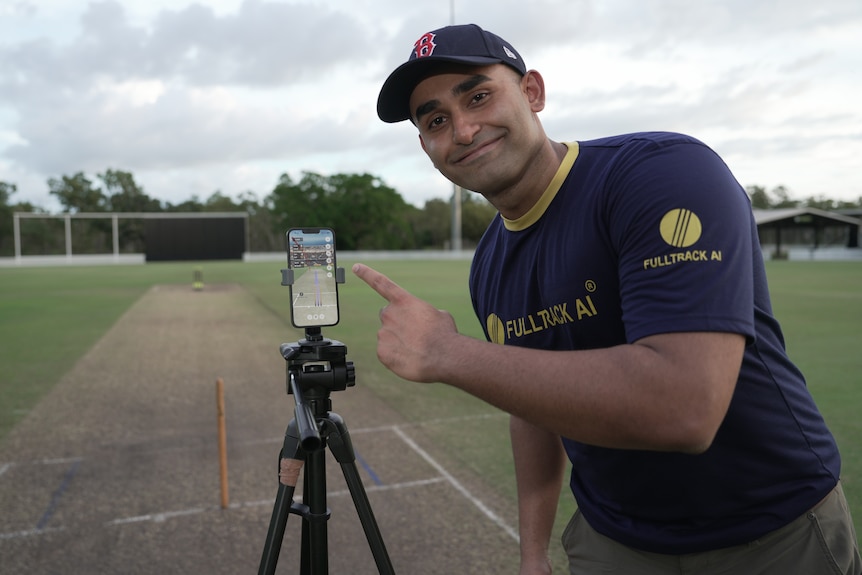 A man smiles at the camera while pointing at a smartphone, which is attached to a tripod and faces a cricket pitch.