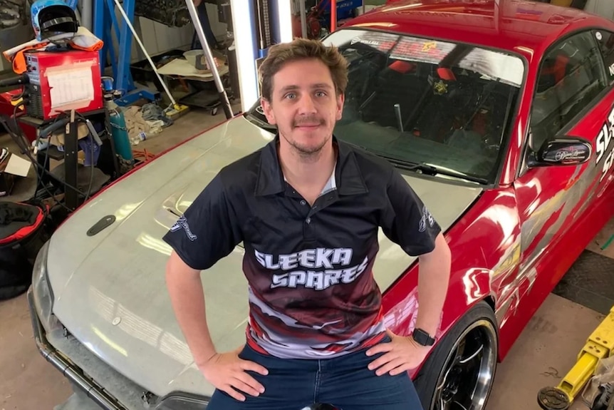 A smiling, dark-haired man sits on a race car in a garage.