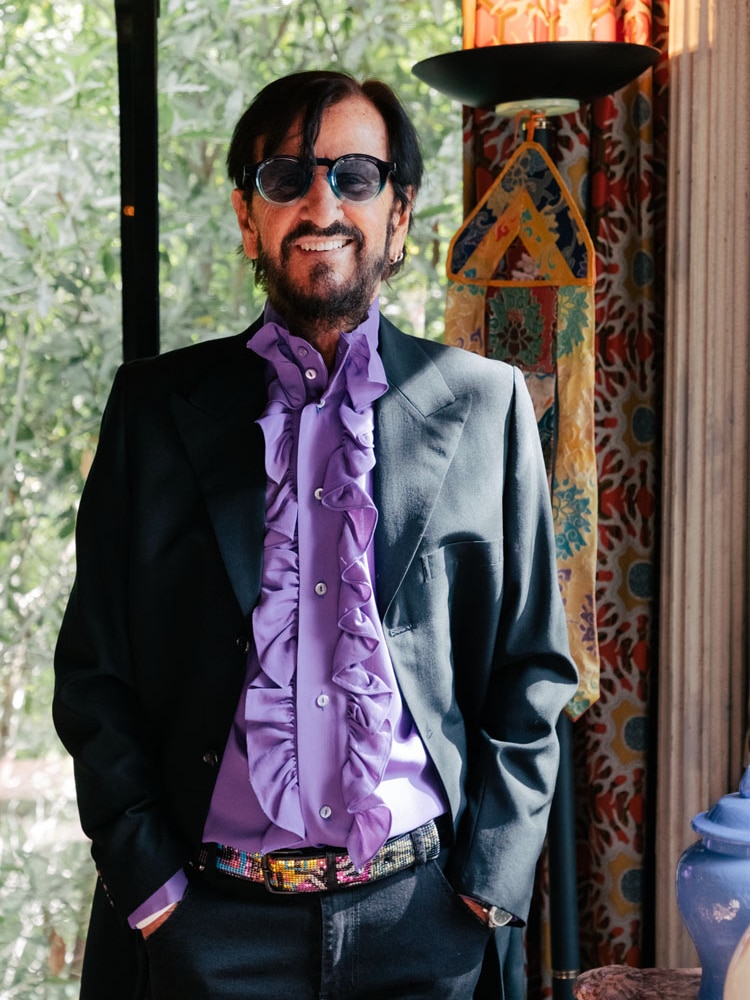 Ringo Starr stands up smiling with hands in pockets. He wears a purple shirt, dark jacket and sunglasses.