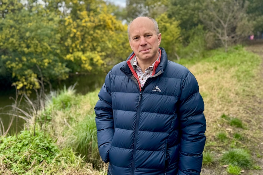Nick Williams wearing a puffer jacket standing in front of a creek with greenery around.