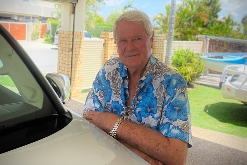 An older man leans on a car to pose for a photo wearing a blue flowery shirt.