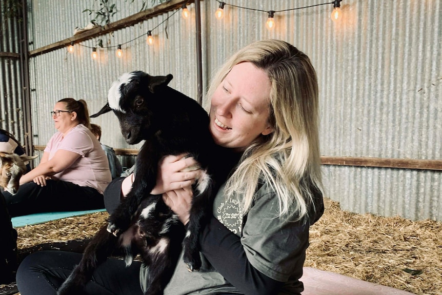 Blonde woman smiling while patting goat, on the floor of a barn