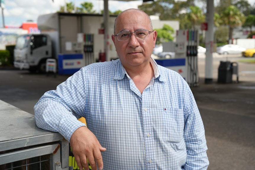A man in a checkered shirt leans agrainst a metal box at a petrol station.