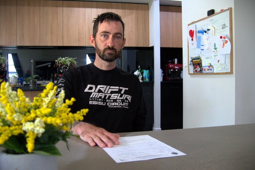A thin middle aged man with dark hair and a somber expression sitting at a kitchen table.