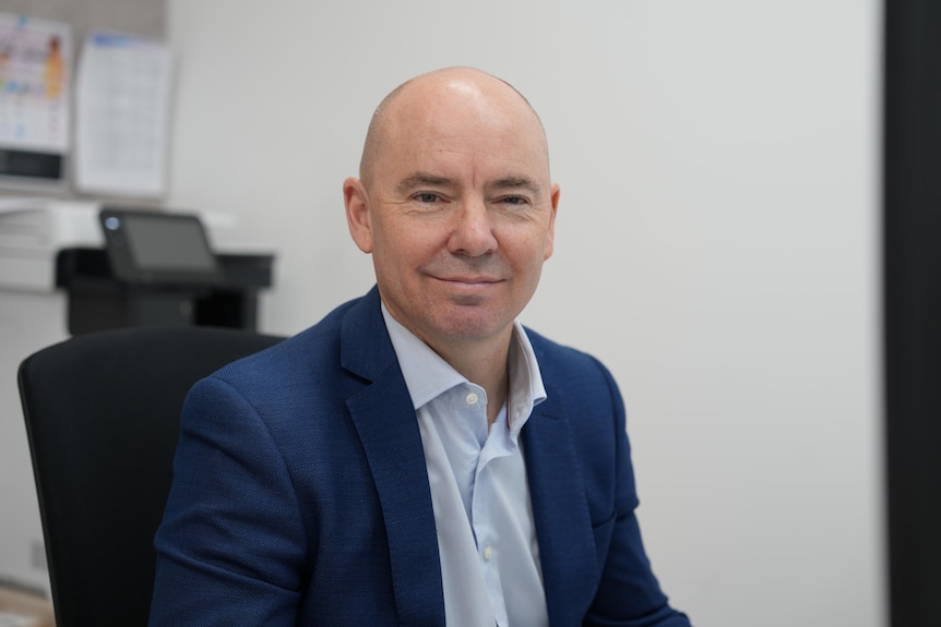 A bald caucasian man wearing a blue suit jacket and shirt smiles