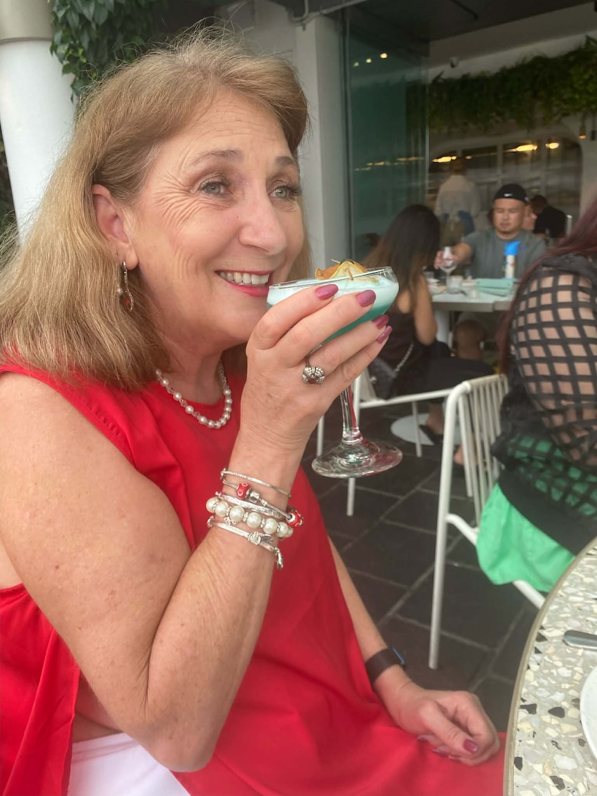 A woman smiles as she holds up a drink while sat outside at a cafe.
