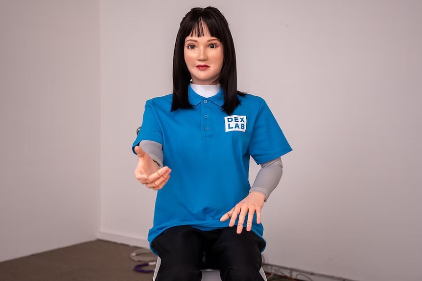 A humanoid robot that appears as a brunette female, offering its right hand while wearing a blue 'DexLab' shirt.