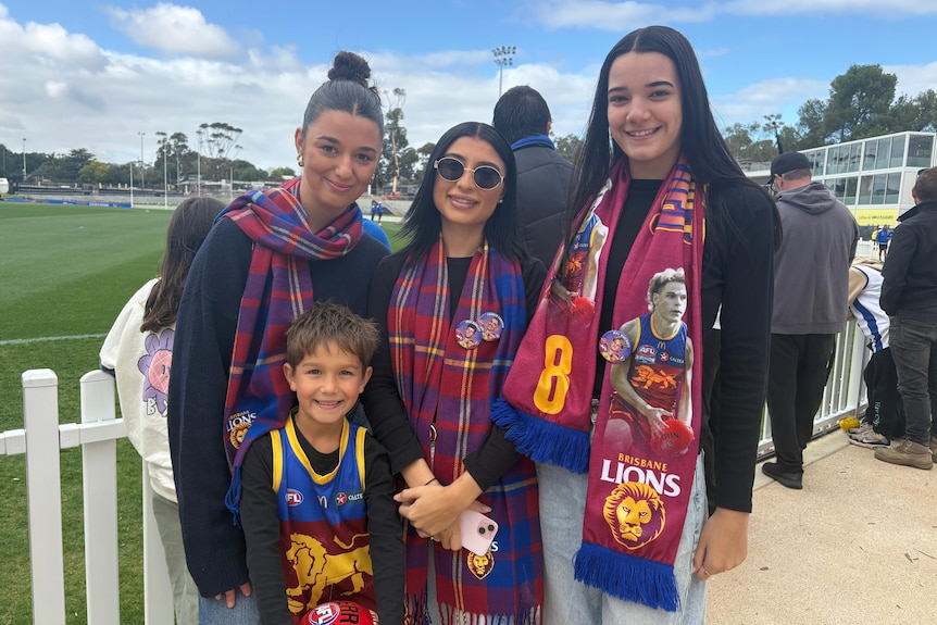 Three young women and a boy wearing Brisbane ions gear next to a white picket fence