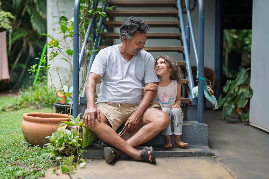 A tan man, brown / gray hair, sitting on stairs outside home, with a young girl (maybe 5) holding his arm, smiling up at him