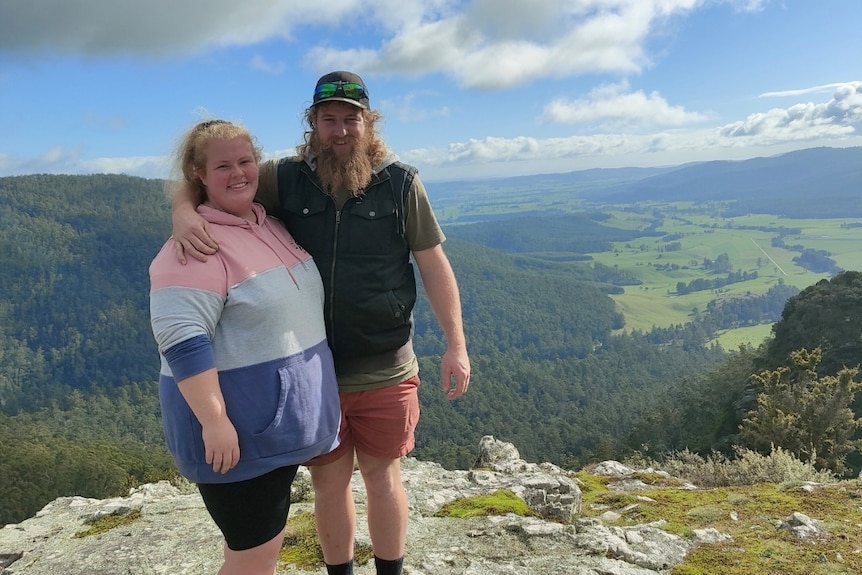A man and woman in athletic gear pose on a cliff overlooking a valley.