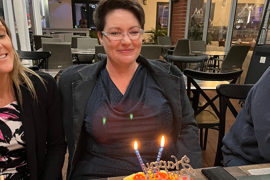 A smiling young woman with short dark hair and glasses sits at a restaurant table in front of a birthday cake.