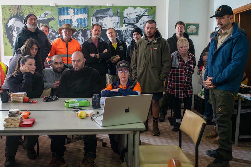 A large group of people huddle around a laptop, serious facial expressions