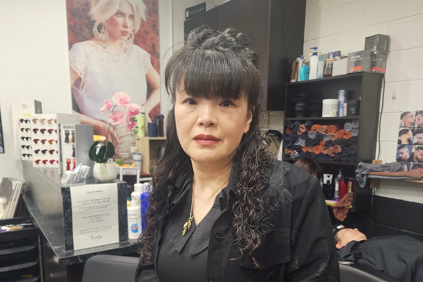 A woman looks directly at the camera as she stands in a hardressing salon.