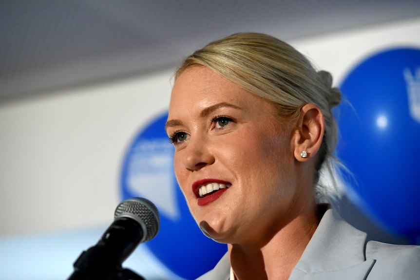 Ashton Hurn stands behind a microphone, with blue balloons behind her