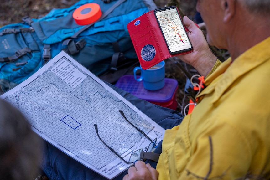 Partial view of a man in a yellow jacket holding a mobile phone while kneeling with a white paper map. Glasses in his hand