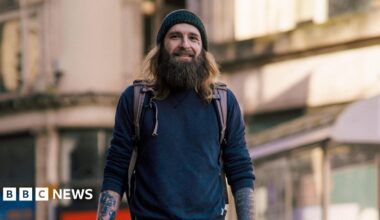 Tom Wilkins stood in Cardiff high street. He has a brown long beard, brown long hair and wears a knitted grey hat. Straps from his backpack can be seen on his shoulder, and he wears a jumper rolled up to his elbows