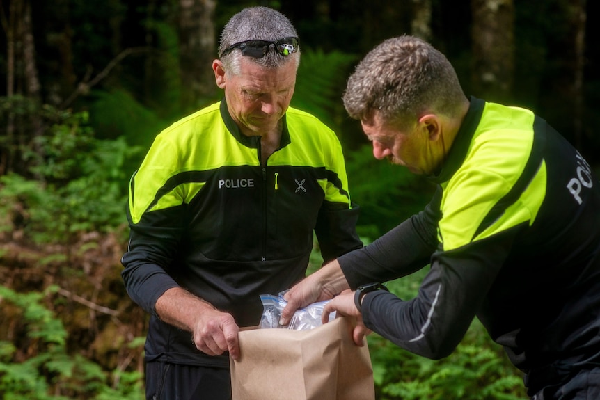 Two police officers in yellow and black hi-vis long-sleeved shirts stand. One holds a brown bag open the other places an item