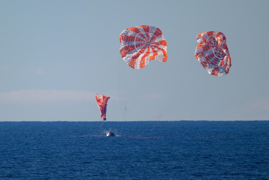 Three white and red parachutes fall down as the spacecraft they're carrying lands in the ocean